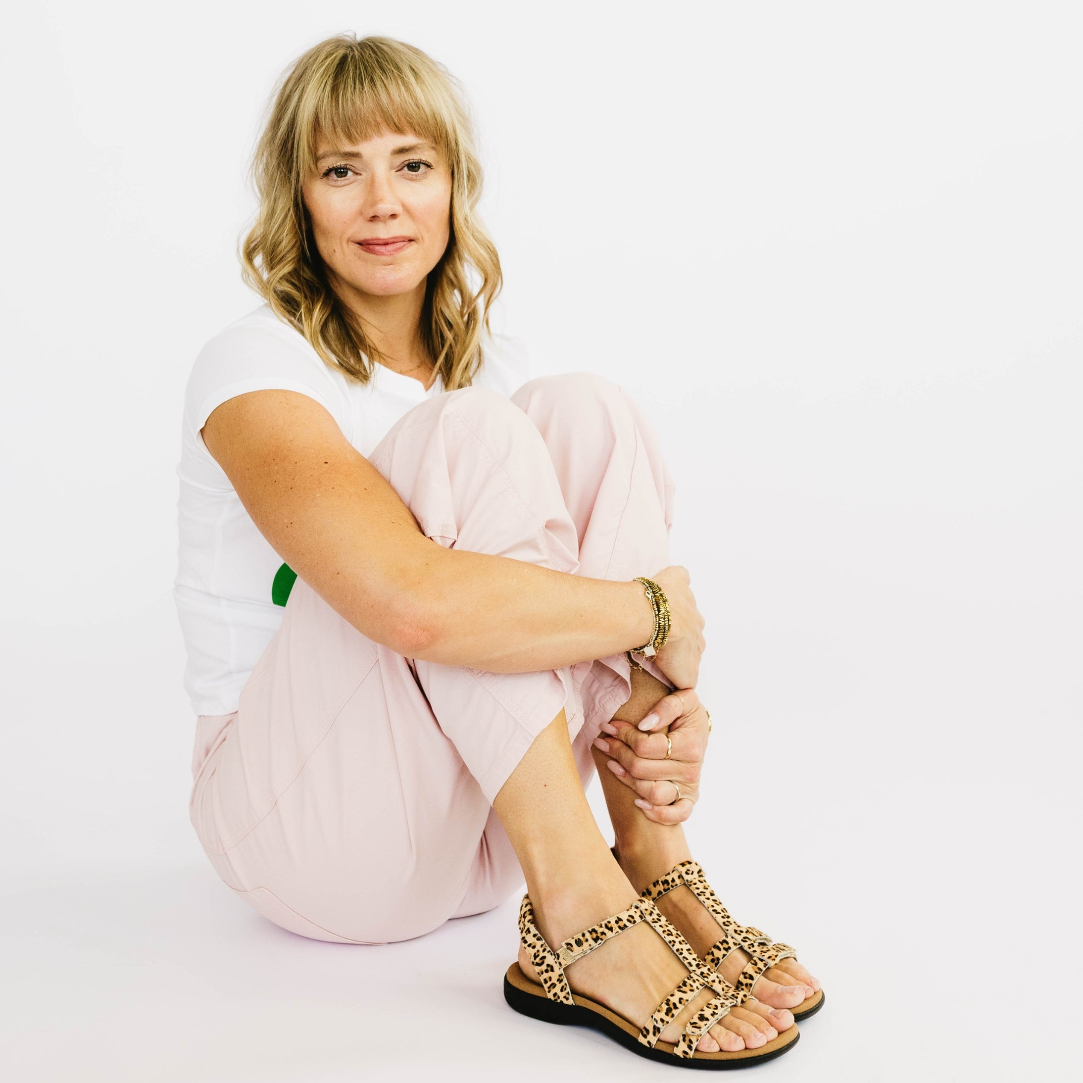 Woman sitting on a white background wearing a white shirt, pink pants, and leopard print sandals.