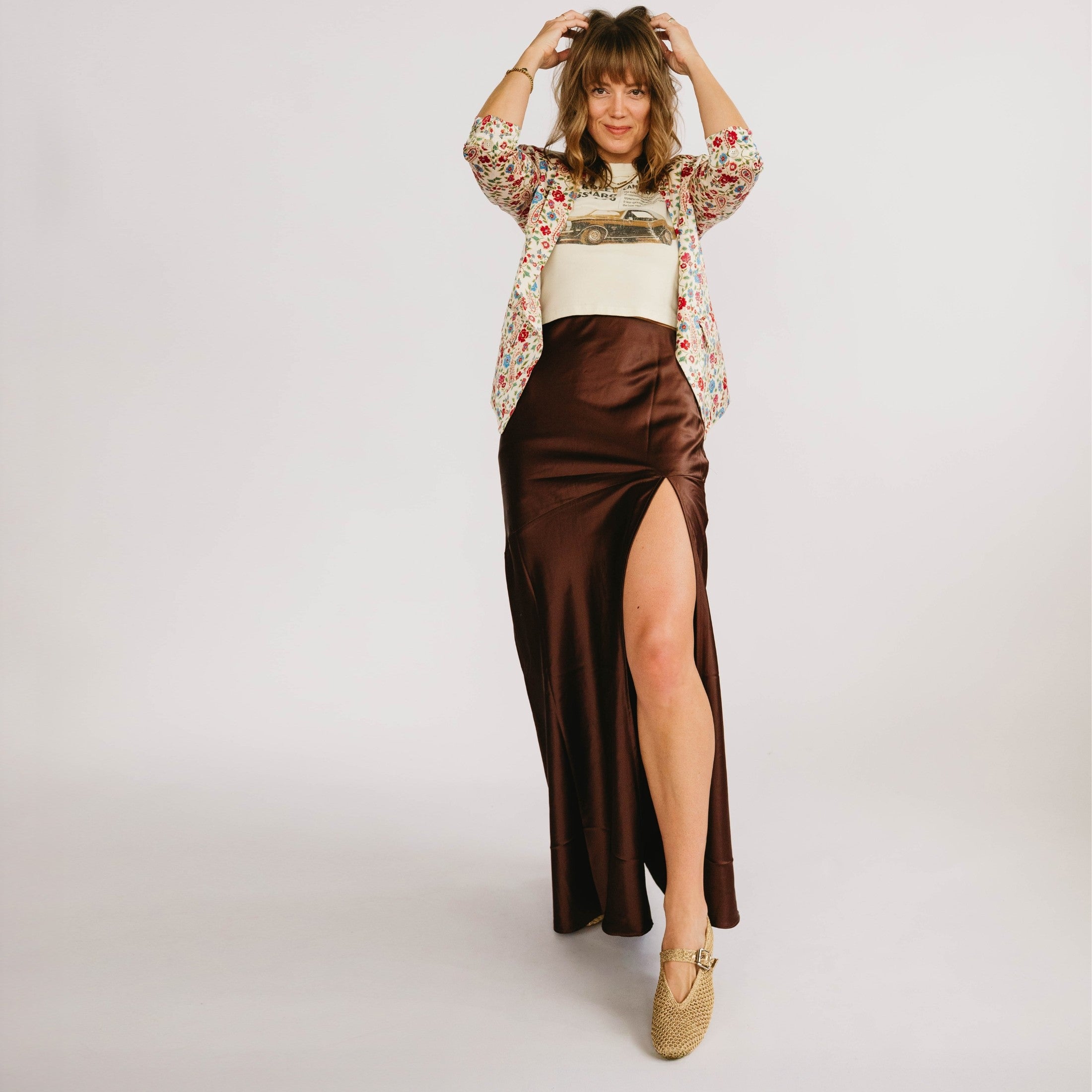 Woman wearing a floral blouse, white top, and brown satin skirt with a slit on a light gray background
