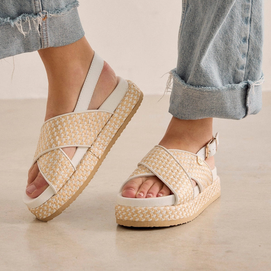 A pair of women's platform sandals with woven beige and white straps, shown on a model's feet with cuffed light-wash jeans against a neutral beige background.