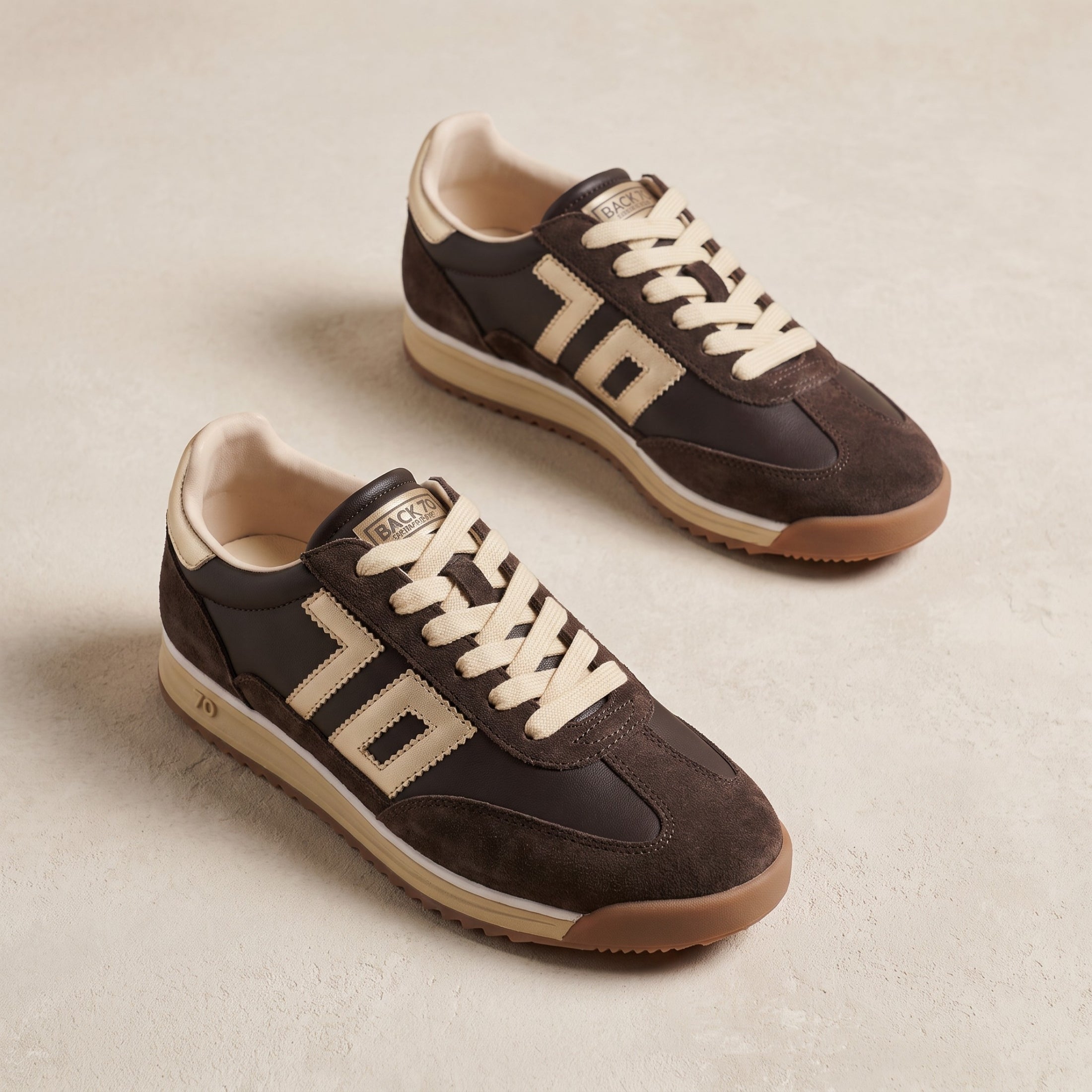 A pair of vintage-style low-top sneakers by Riddell, featuring dark brown suede and leather panels, cream-colored laces and numerals, and a tan rubber sole, displayed on a light beige textured surface.